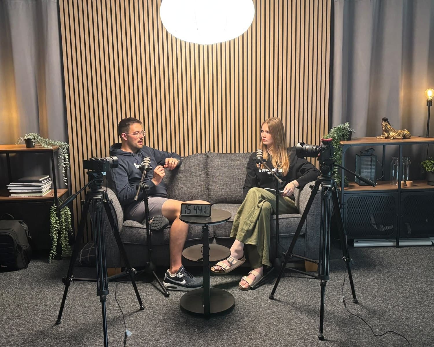 Two people recording on the sofa set, filmed from two camera positions on tripods.
