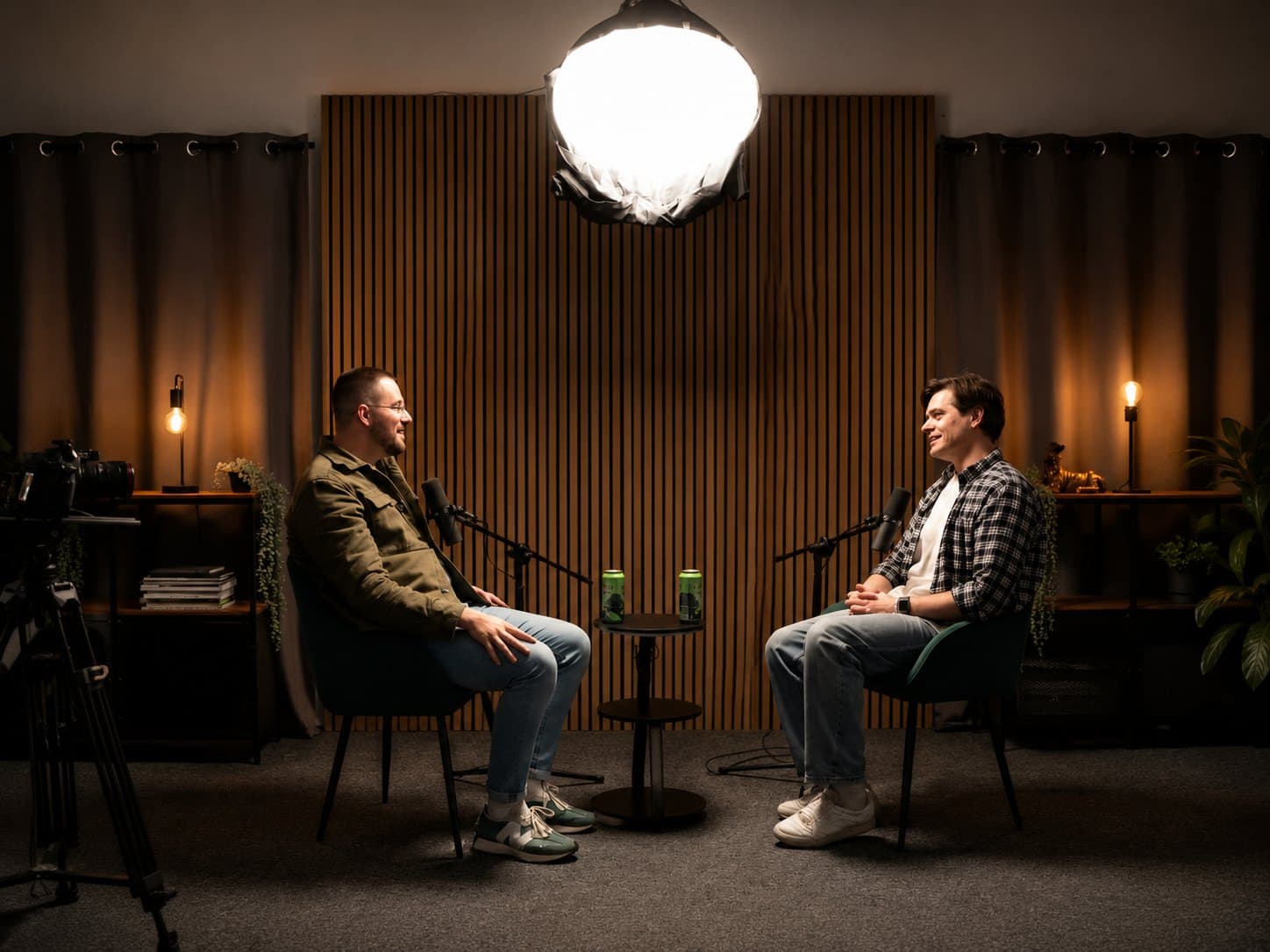 Two people recording a podcast in the Lincoln Podcast Studio, seated facing each other with microphones, under a large softbox light against warm wood panelling.